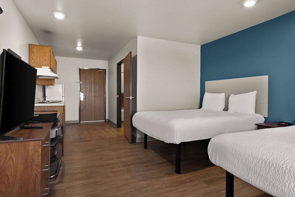A hotel room with a bed, a desk with a television, and a kitchenette.