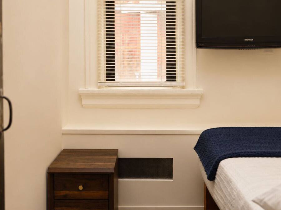 Un dormitorio con cama, una mesita de noche de madera, ventana con persianas y televisión montada en la pared.
