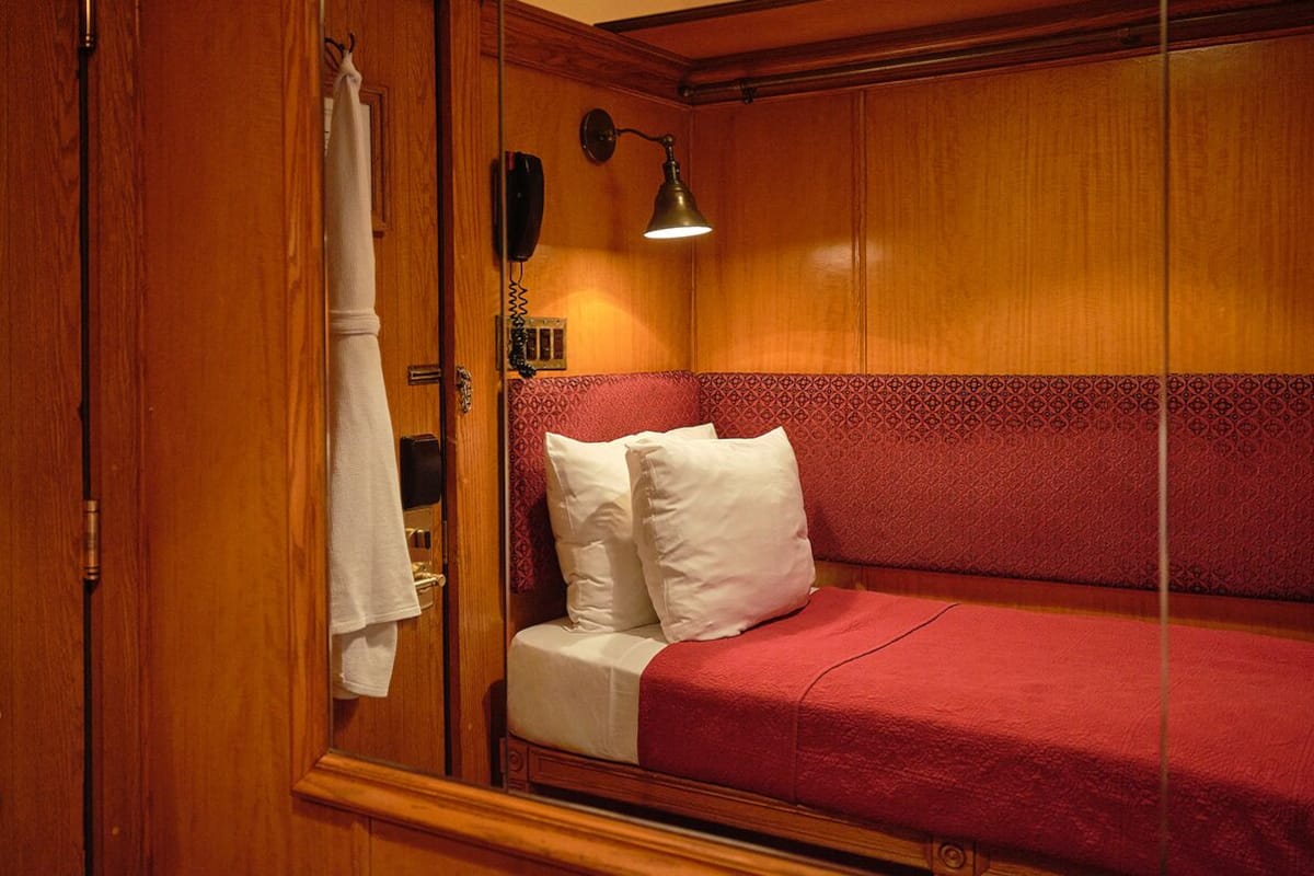 A wooden room with a bed, a red headboard, a wall-mounted lamp, and a telephone.