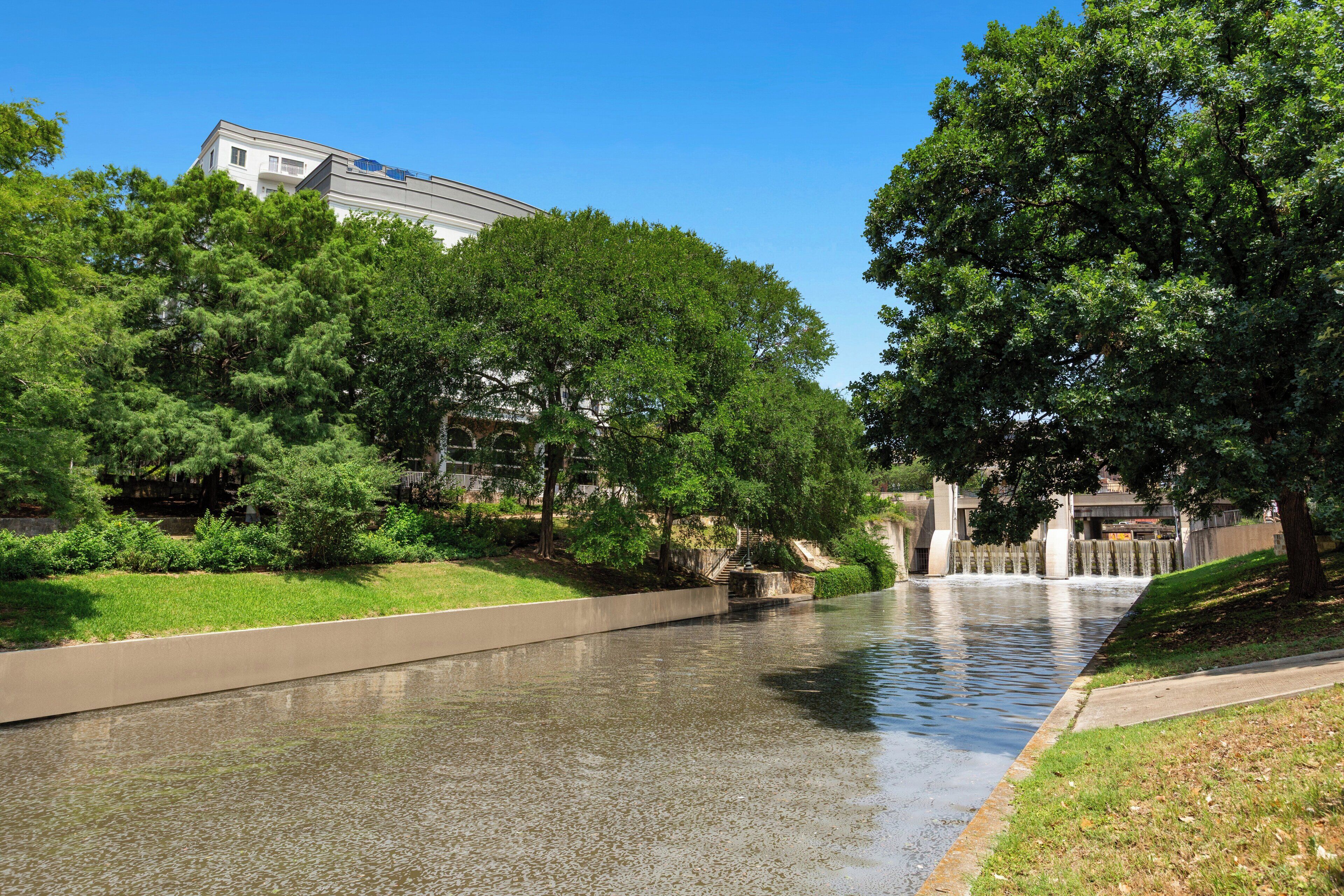 A river with a concrete walkway alongside it, surrounded by lush green trees and a building in the background.