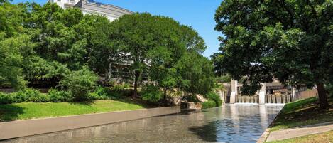 A river with a concrete walkway alongside it, surrounded by lush green trees and a building in the background.