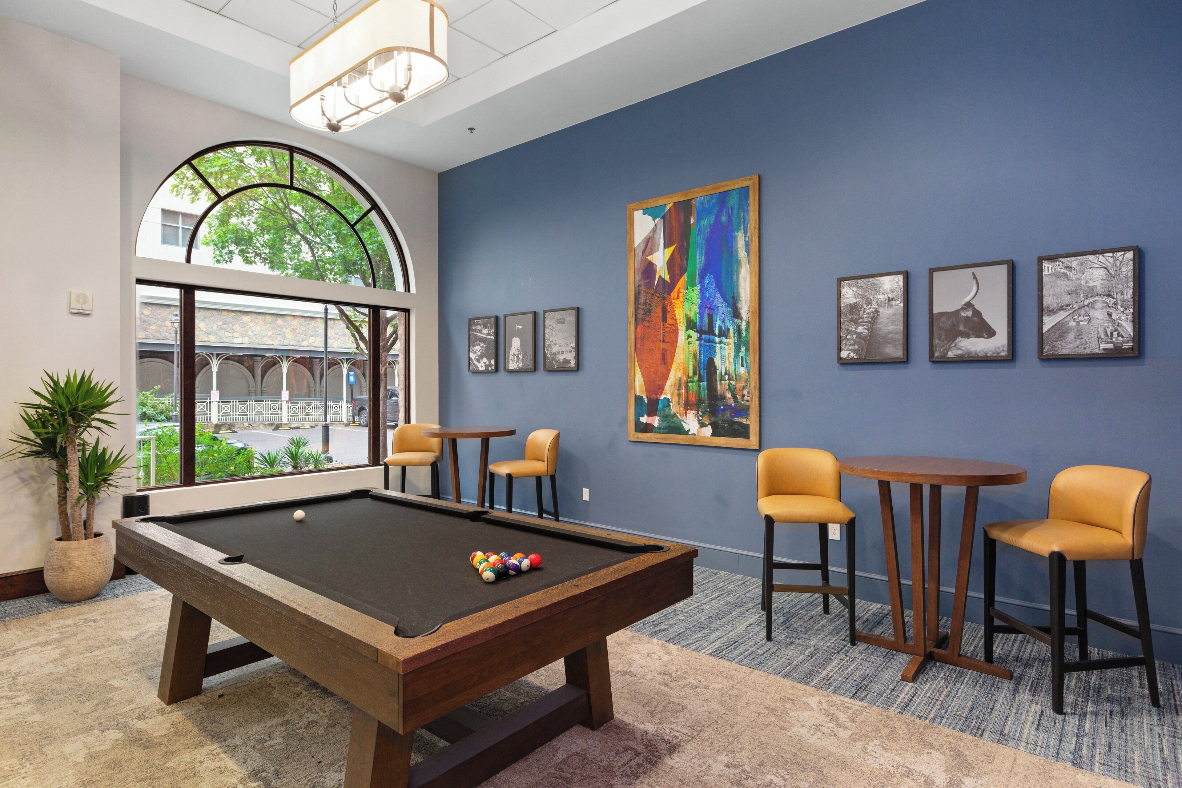 A pool table in a room with a large window, several framed pictures on the wall, and a potted plant.