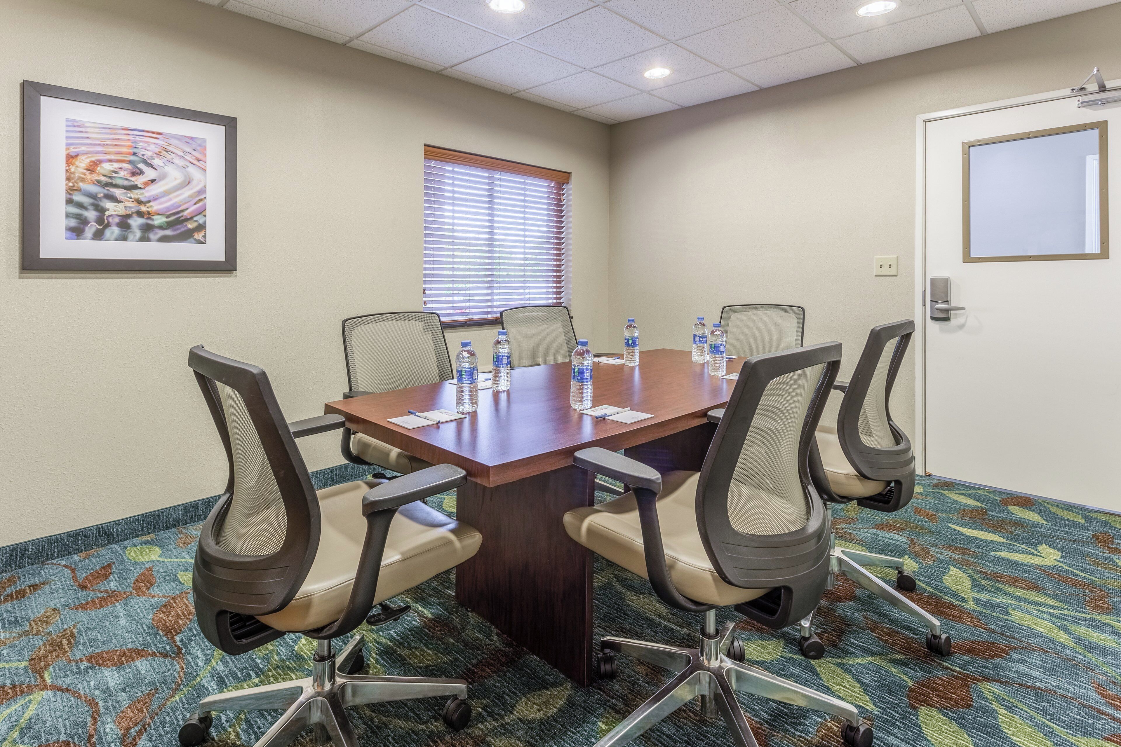 A conference room with a long table, four chairs, water bottles, a framed abstract painting, and a window with blinds.