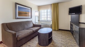 A hotel room with a sofa, a small round table, a flat-screen TV, a window with curtains, and a framed picture on the wall. - Candlewood Suites Aurora-Naperville by IHG (Aurora)