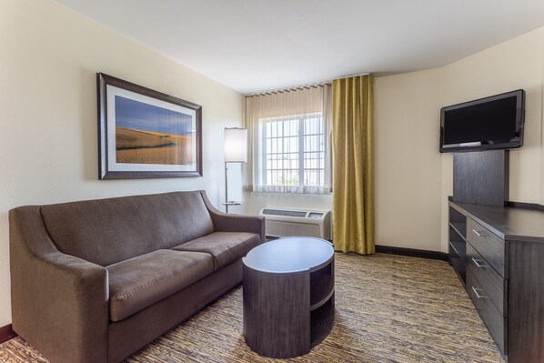 A hotel room with a sofa, a small round table, a flat-screen TV, a window with curtains, and a framed picture on the wall. - Candlewood Suites Aurora-Naperville by IHG (Aurora)