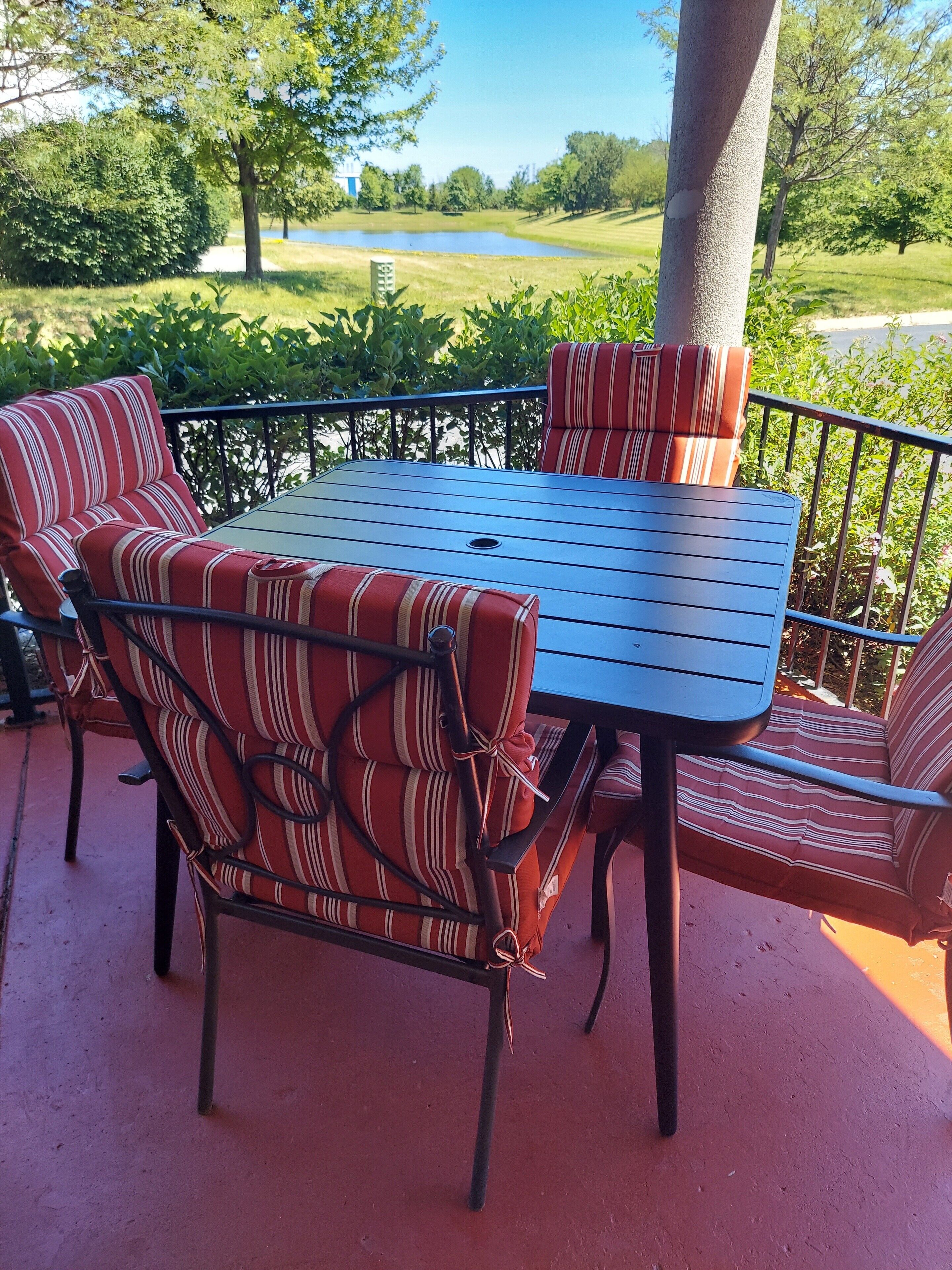 An outdoor patio with a metal table and striped cushioned chairs, surrounded by a metal railing and a view of a golf course.