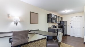 A compact hotel room with a kitchenette, a dining area with a table and chairs, a wall-mounted lamp, and a door. - Candlewood Suites Aurora-Naperville by IHG (Aurora)