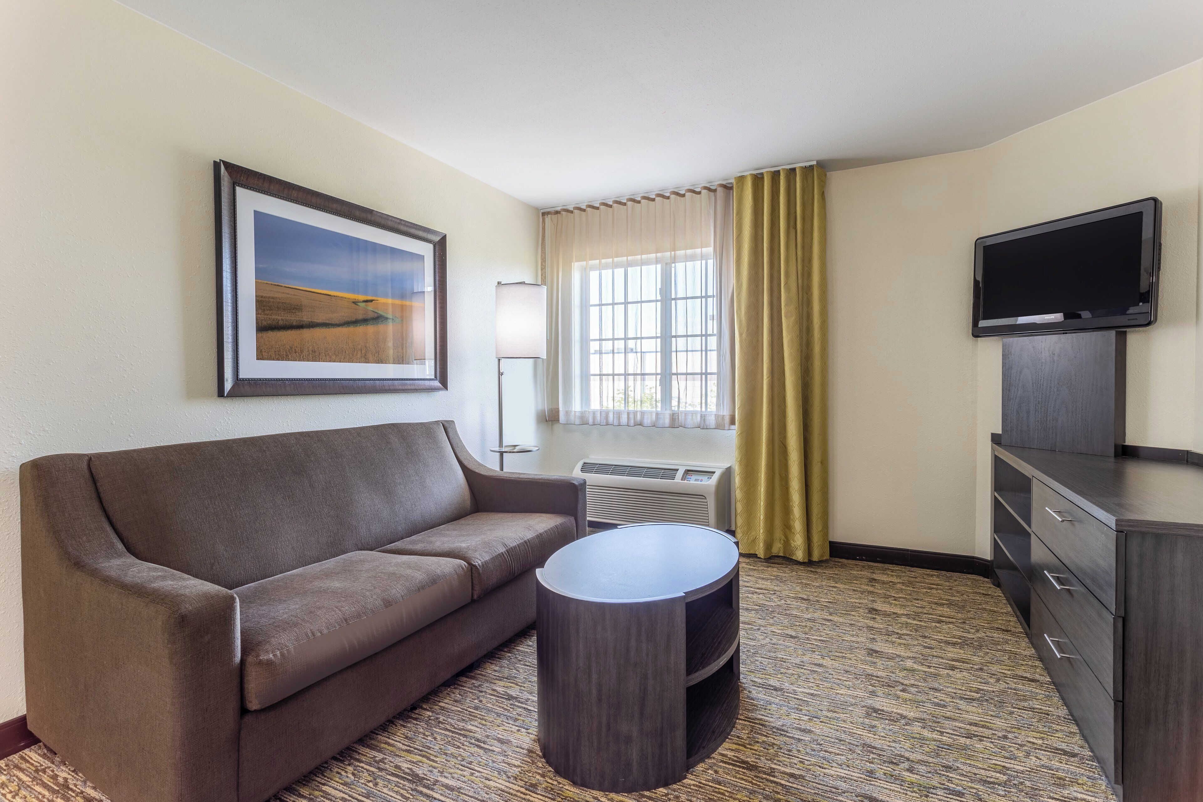 Quarto de hotel com sofá, mesa redonda pequena, televisão e janela com cortinas.