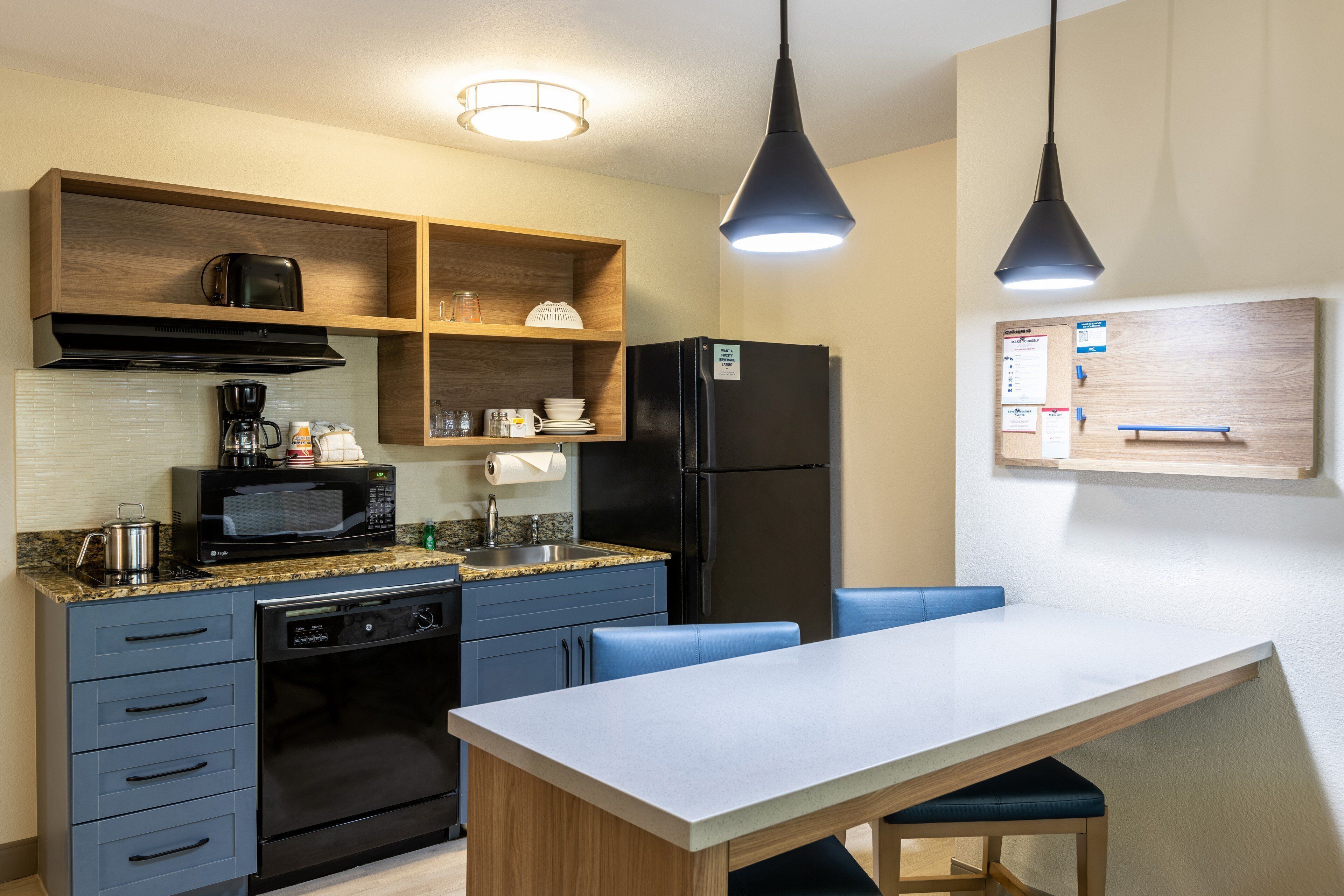 A modern kitchen with a built-in microwave, coffee maker, and a refrigerator. There is a wooden shelf above the sink and a white table with blue chairs.