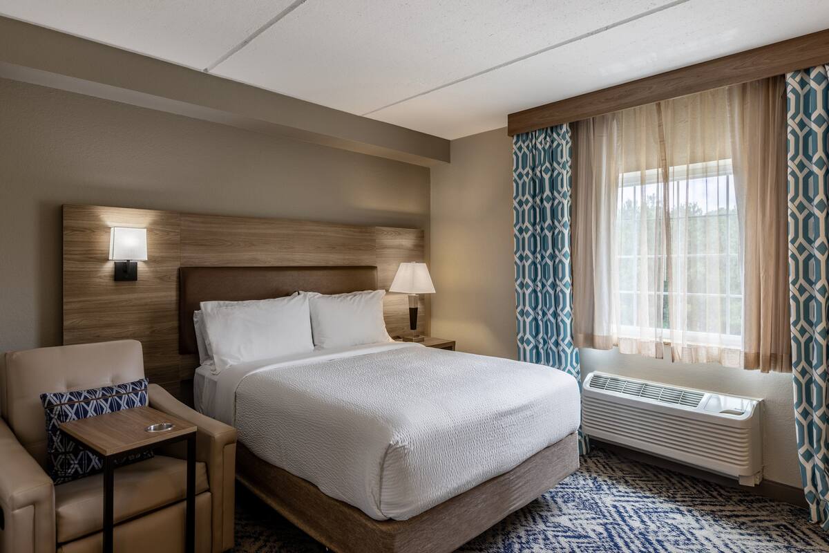 Une chambre d’hôtel avec un grand lit, une table de chevet avec une lampe, une chaise avec un coussin à motifs et une fenêtre avec des rideaux à motifs.
