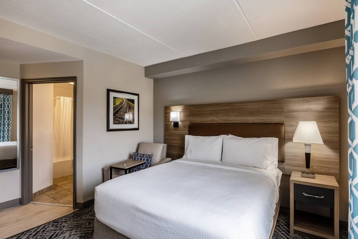 Une chambre d’hôtel avec un grand lit, une table de chevet avec une lampe, une chaise, une petite table et un tableau au mur.