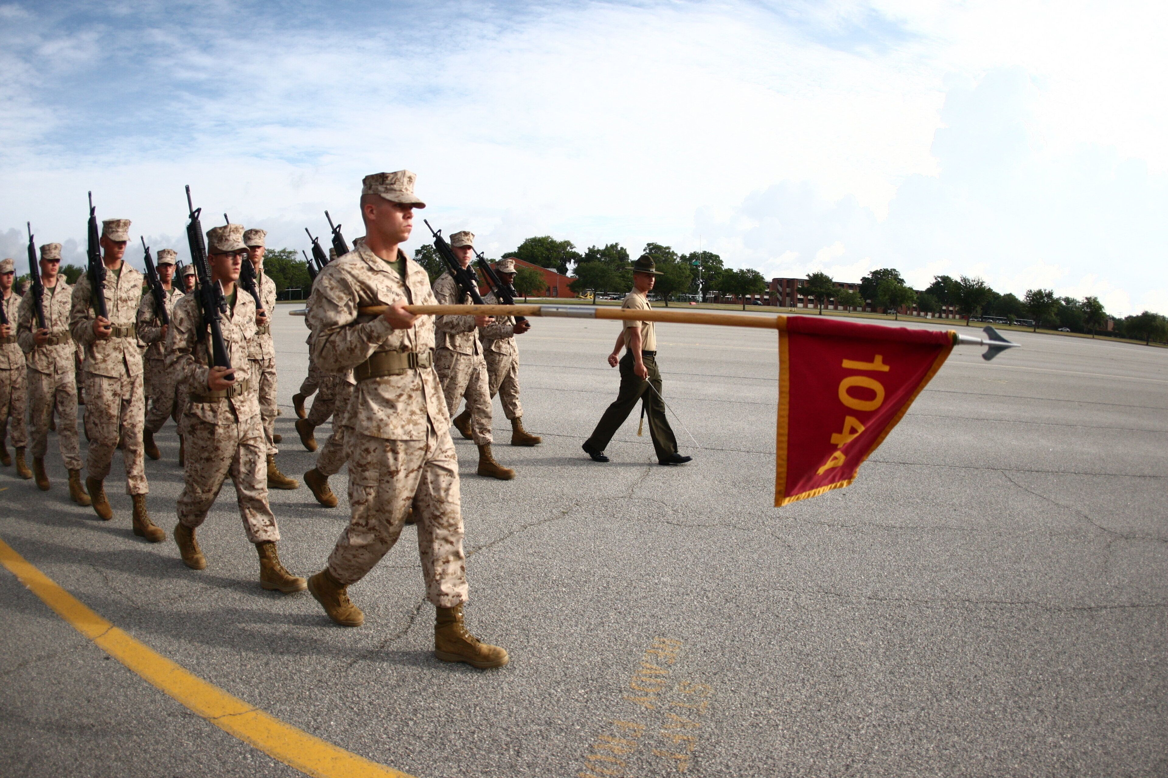 A group of military personnel in uniform marching on a paved surface with a flag being carried.