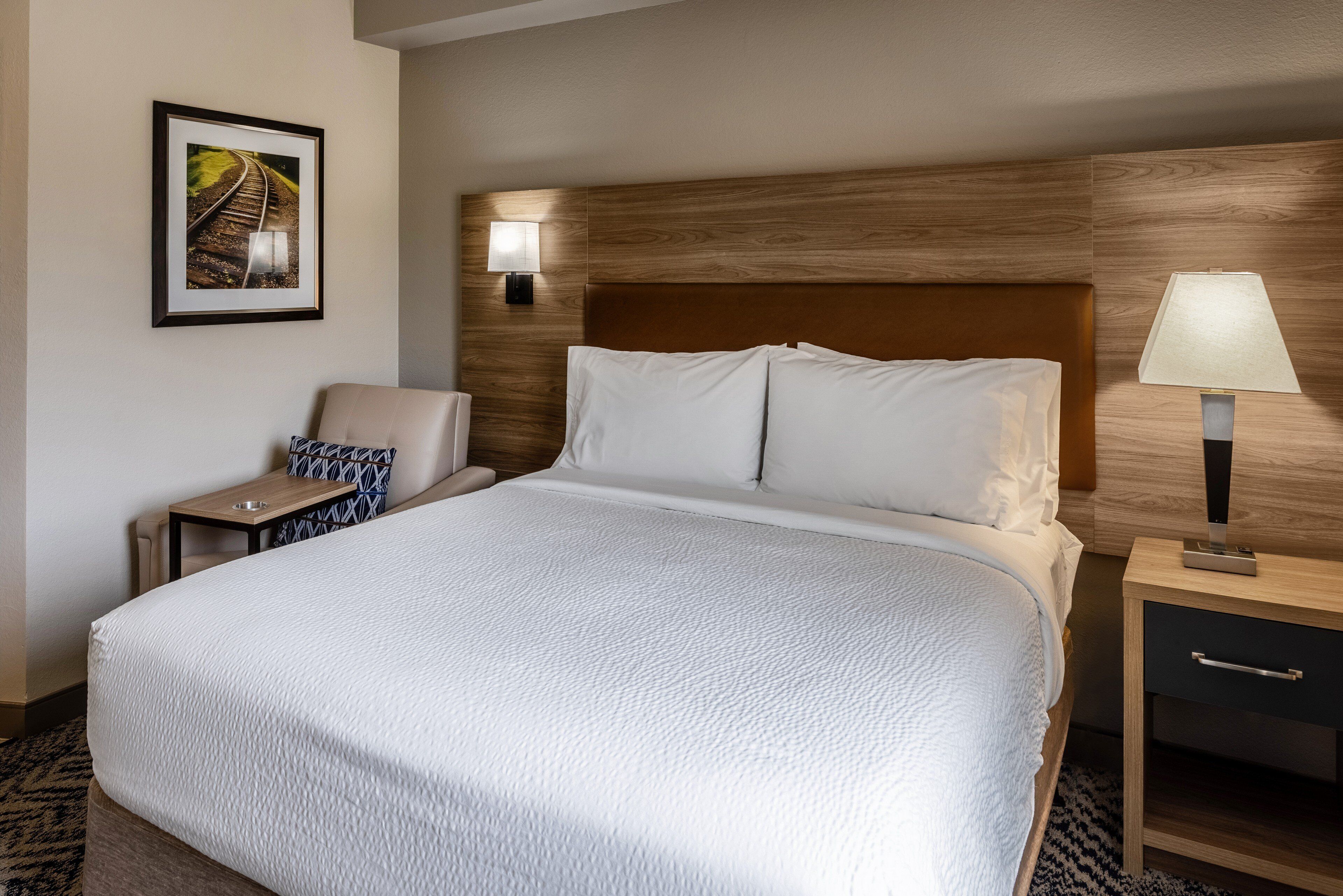 Habitación de hotel con una cama grande, una mesita de noche con lámpara, una silla y un cuadro en la pared.