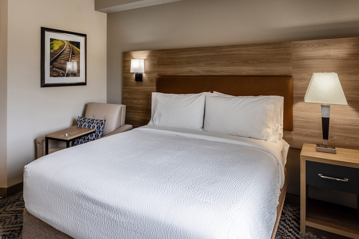 A hotel room with a large bed, a nightstand with a lamp, a chair, a small table, and a framed picture on the wall.