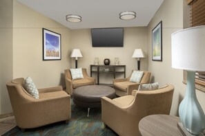 Ein Hotelzimmer mit beigen Wänden, einem Flachbildfernseher über einem kleinen Tisch, zwei Sessel mit gemusterten Kissen, einem runden Hocker und einer großen Stehlampe.