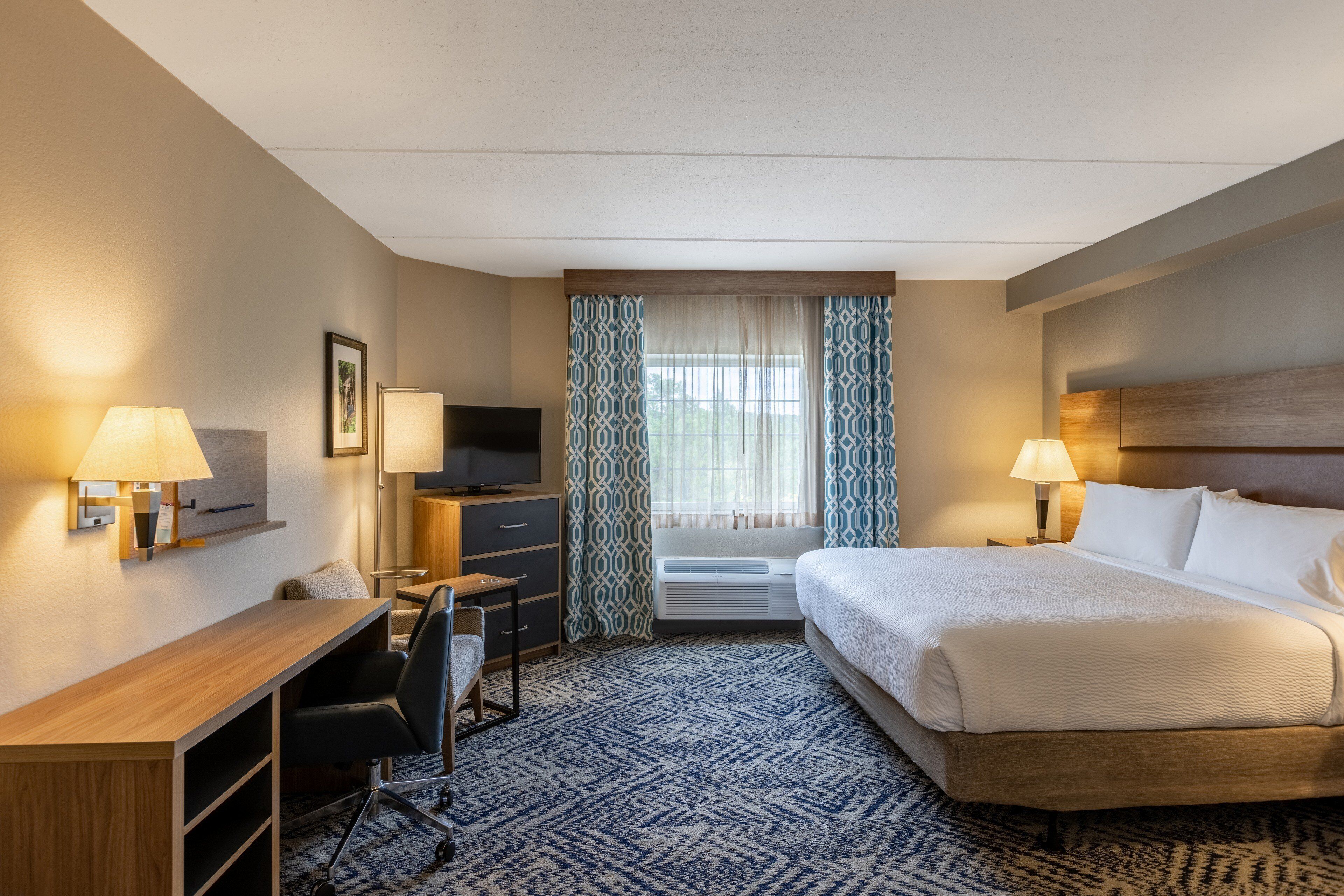 Une chambre d’hôtel avec un lit, un bureau, une chaise, une télévision et une fenêtre avec des rideaux.