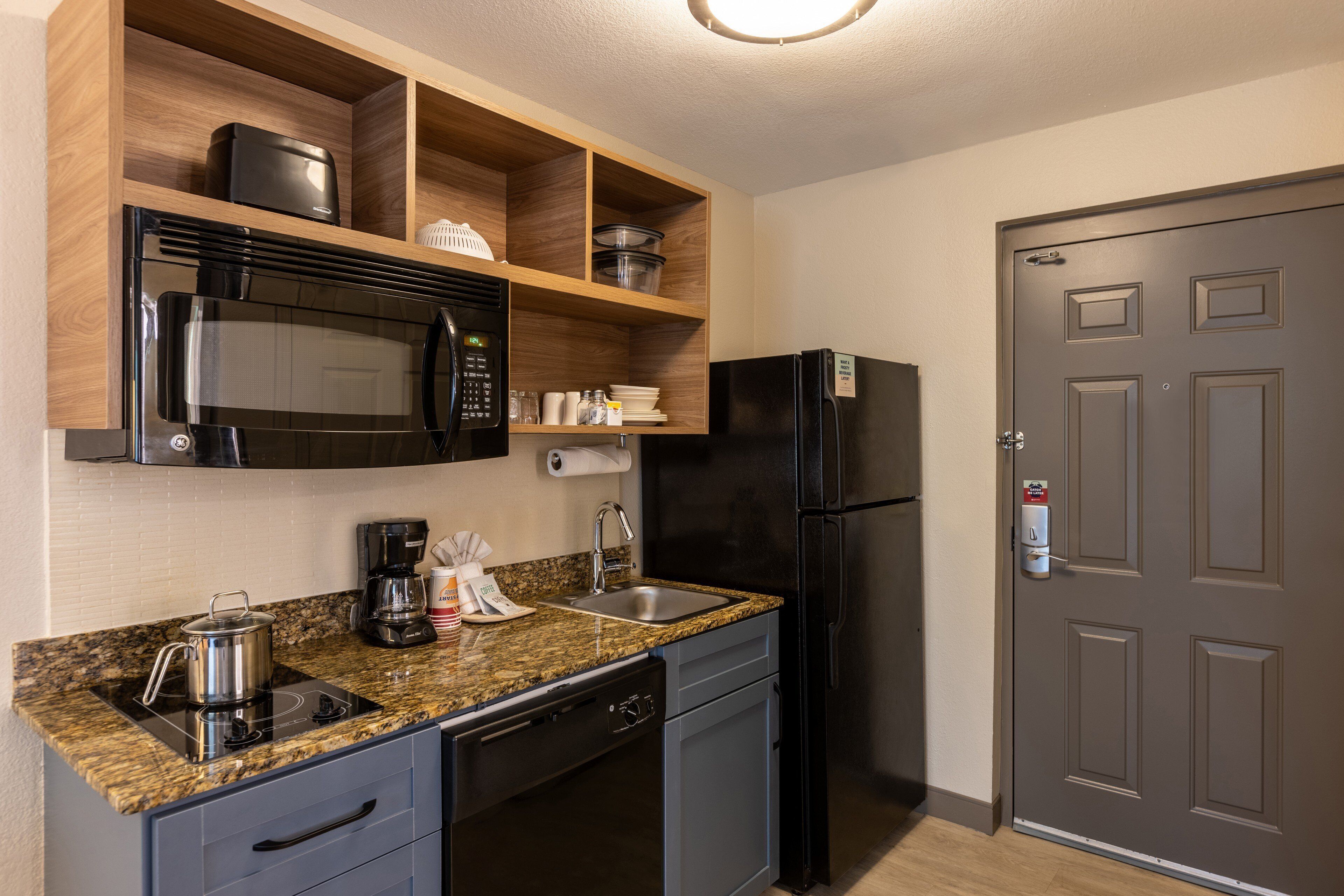 Cocina compacta con microondas, estufa, pileta y refrigerador. Hay repisas de madera sobre la mesada y una puerta a la derecha.