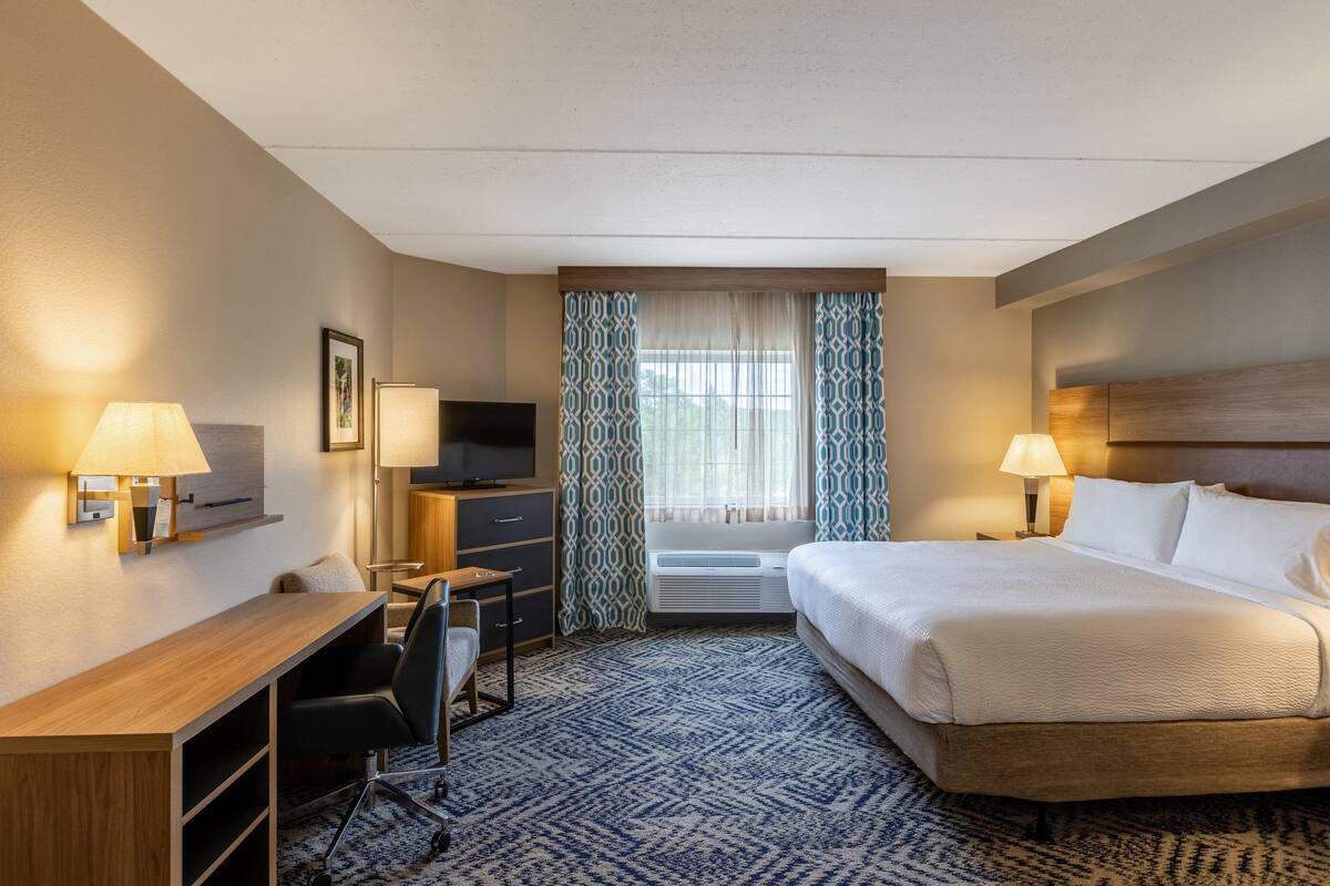 Une chambre d’hôtel avec un lit, un bureau, une chaise, une télévision et une fenêtre avec des rideaux.