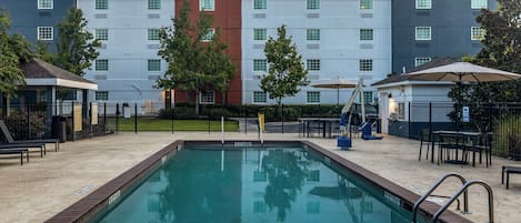Une piscine bordée de briques, entourée d’un espace de détente extérieur et, en arrière-plan, un bâtiment doté de nombreuses fenêtres.
