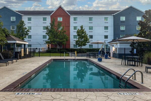 Ein Pool mit Backsteinrand, umgeben von Außenmöbeln und einem Gebäude mit mehreren Fenstern im Hintergrund.