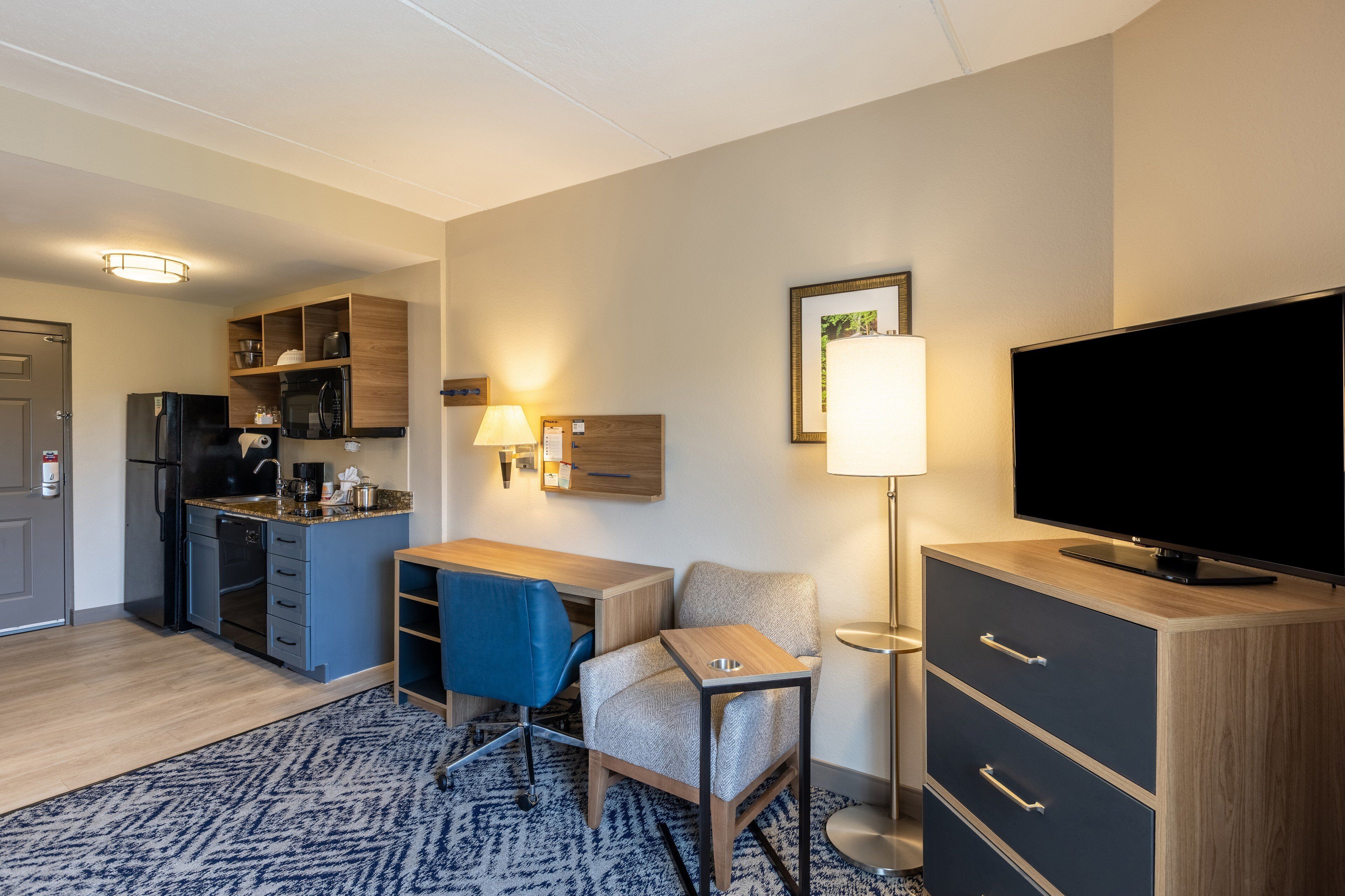 Una habitación de hotel compacta con kitchenette, televisor de pantalla plana, escritorio con silla y alfombra estampada.