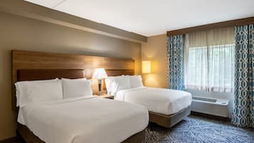 A hotel room with two beds, a nightstand with a lamp, a window with curtains, and a wall-mounted air conditioning unit.