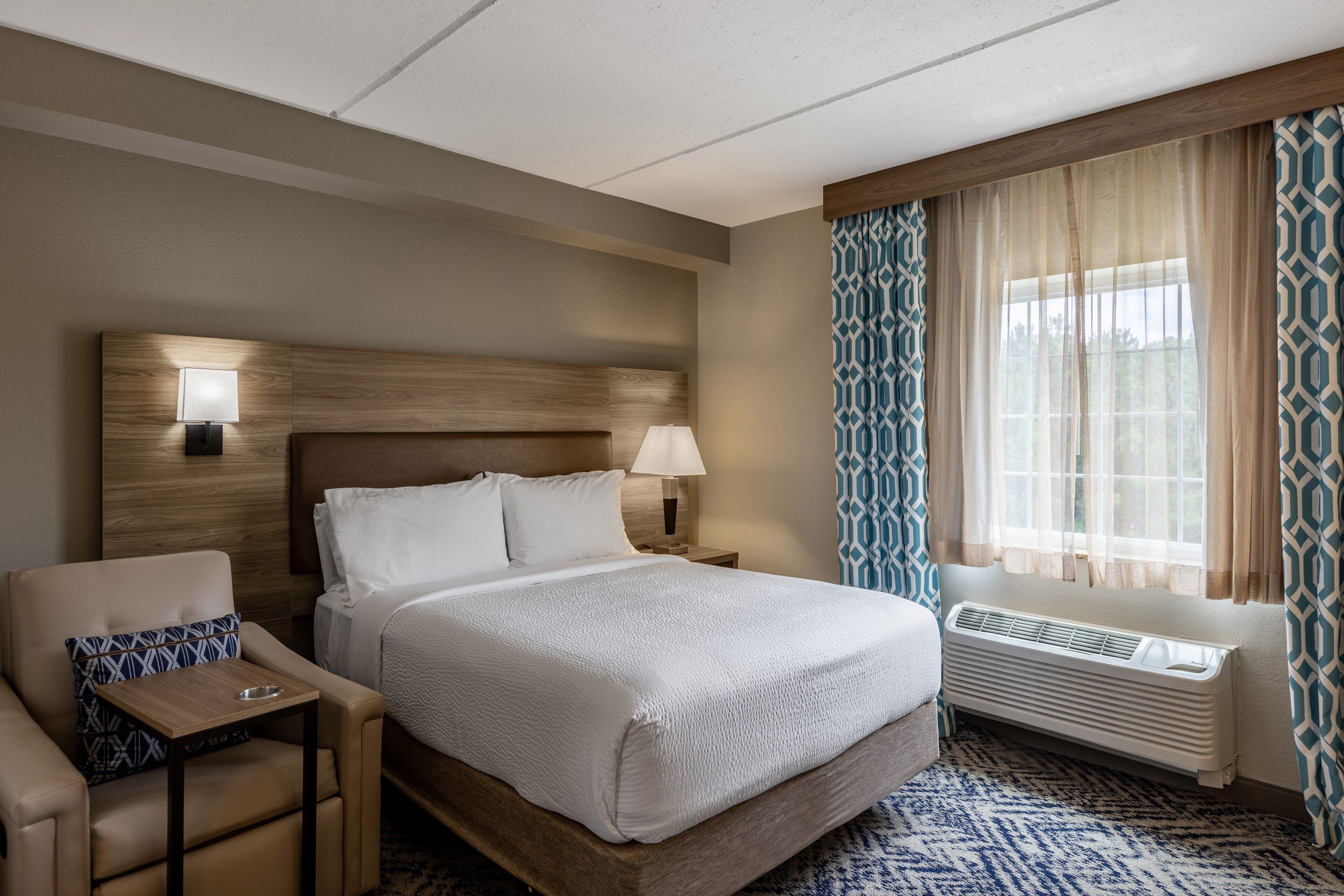 A hotel room with a bed, a chair, a nightstand, a lamp, a window with curtains, and a radiator.