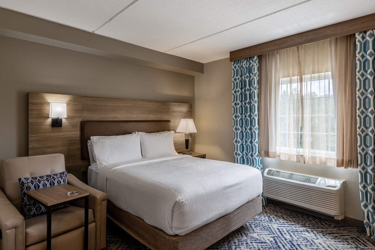 A hotel room with a large bed, a bedside table with a lamp, a chair with a patterned cushion, and a window with patterned curtains.