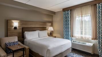 A hotel room with a bed, a chair, a nightstand, a lamp, a window with curtains, and a radiator.