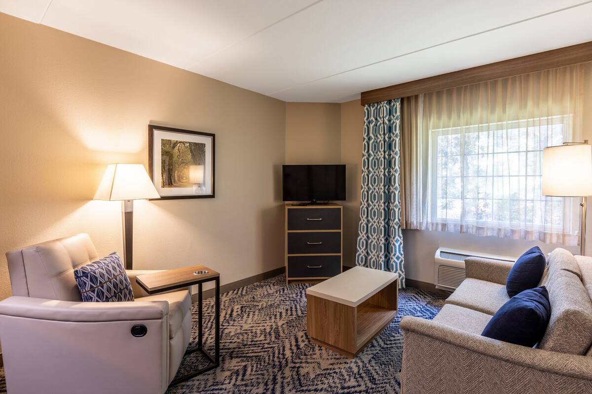 Une chambre d’hôtel avec un canapé, un fauteuil, une petite table, une télévision, une armoire et une fenêtre avec des rideaux.