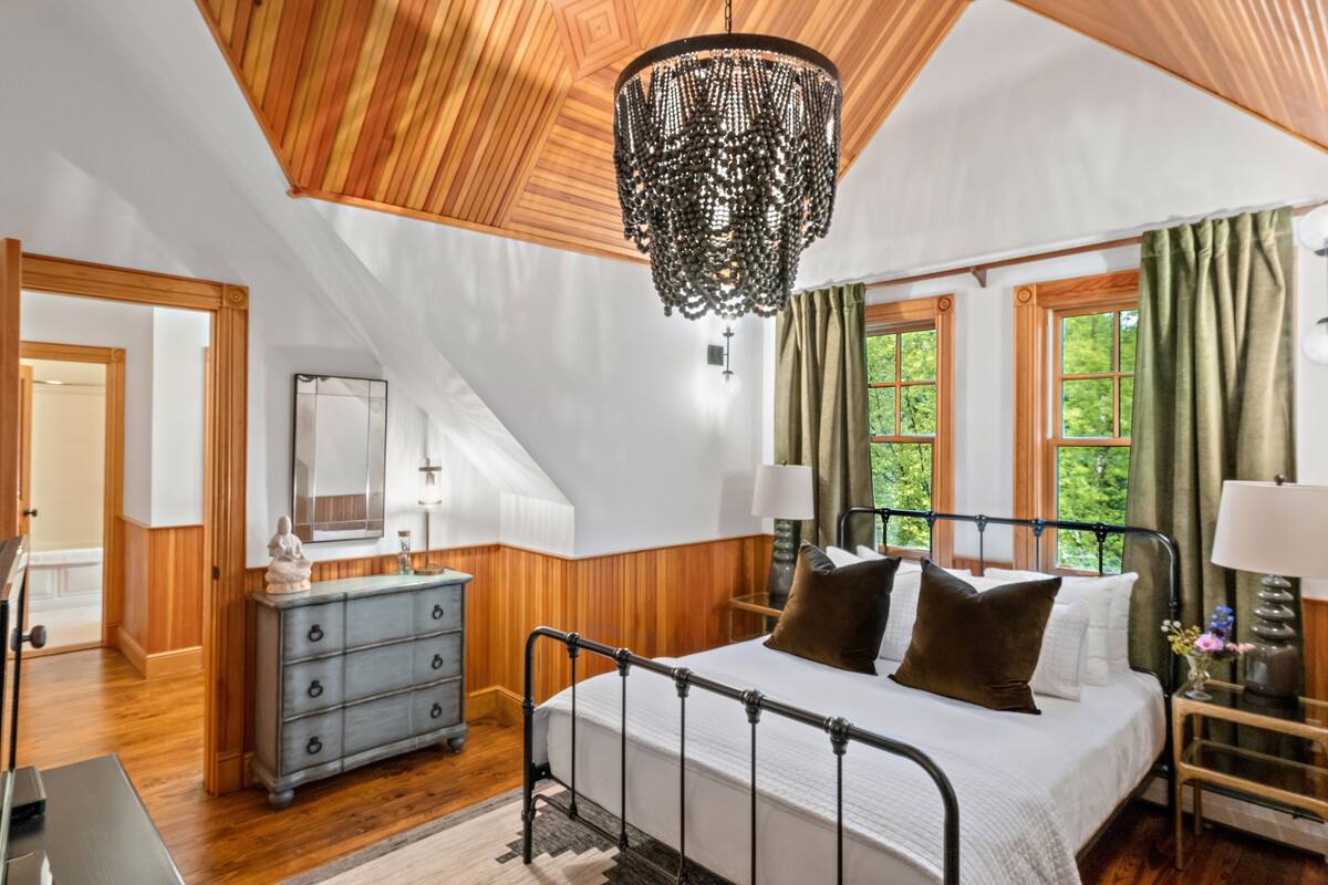 A bedroom with a wooden ceiling, a large bed with a metal frame, a dresser, and a chandelier.