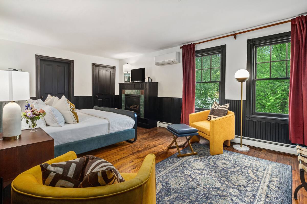 A bedroom with a bed, a yellow armchair, a blue ottoman, a floor lamp, and a large window with red curtains.