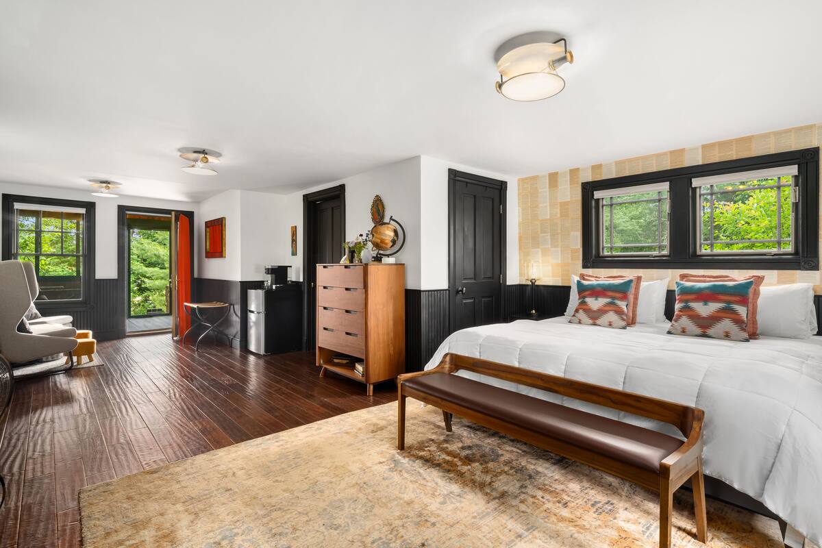 A bedroom with a large bed, a wooden bench, a dresser, and a desk. There are windows with views of greenery outside.