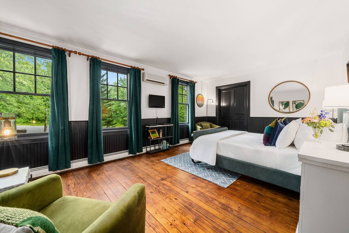 A bedroom with a bed, a green armchair, a wooden floor, and large windows with curtains.