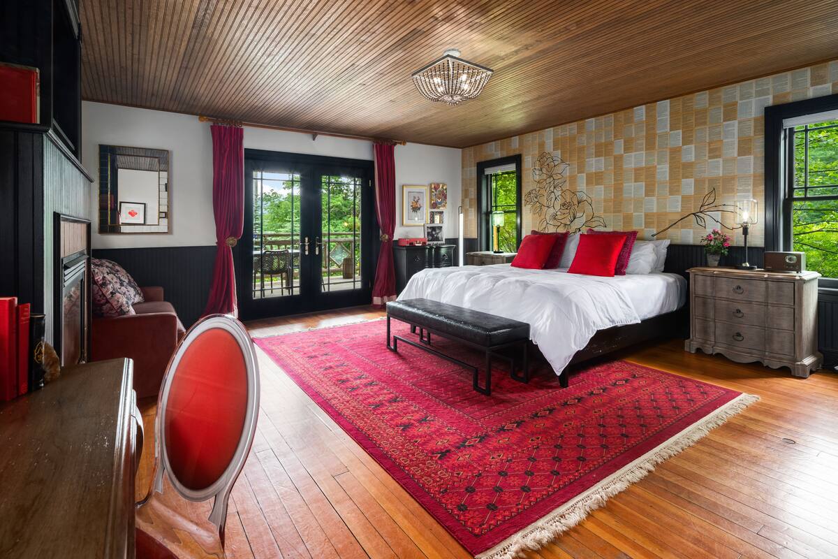 A bedroom with a large bed, a wooden ceiling, a red rug, and a view of greenery outside.