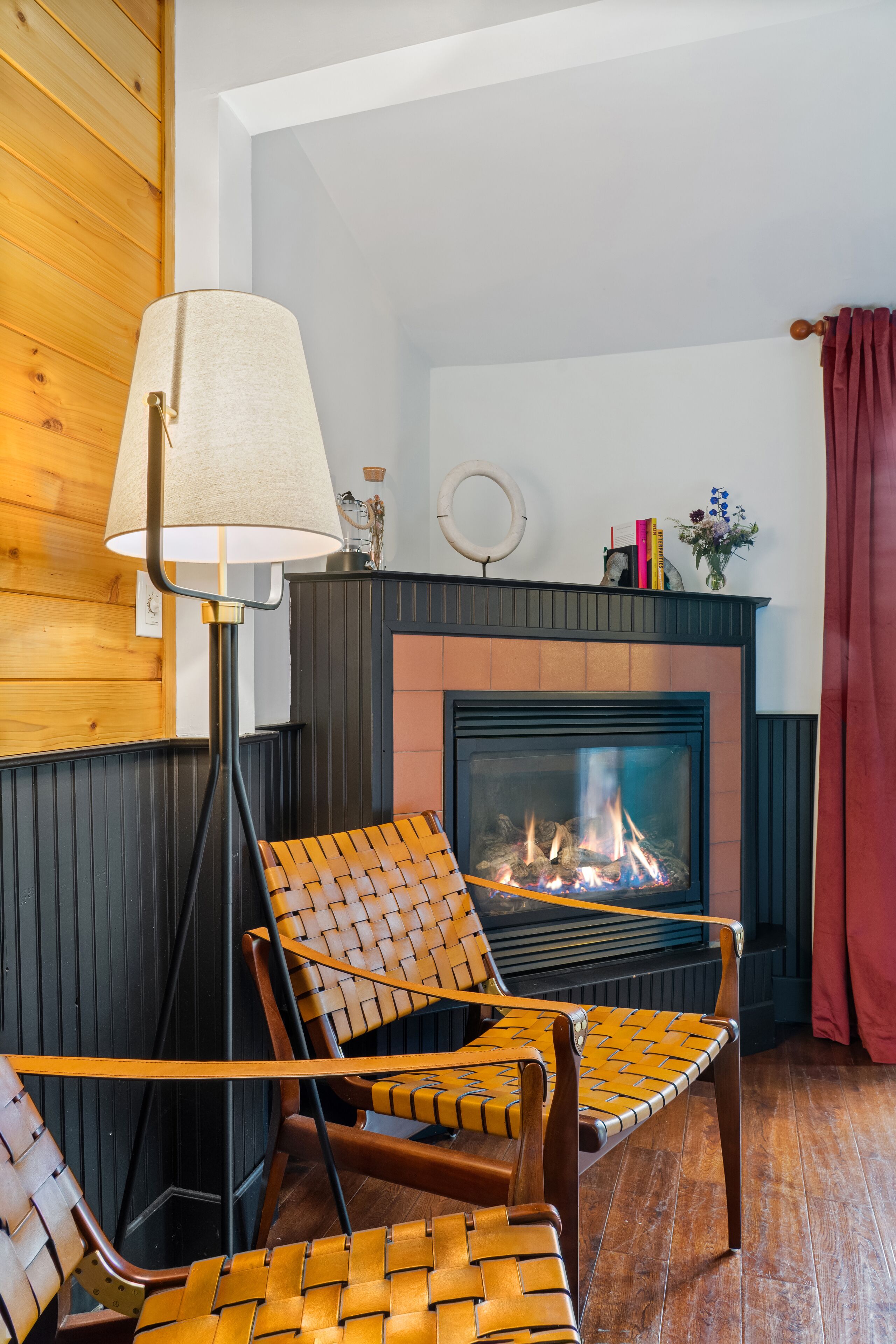 A cozy room with a fireplace, two woven chairs, a floor lamp, and a red curtain.
