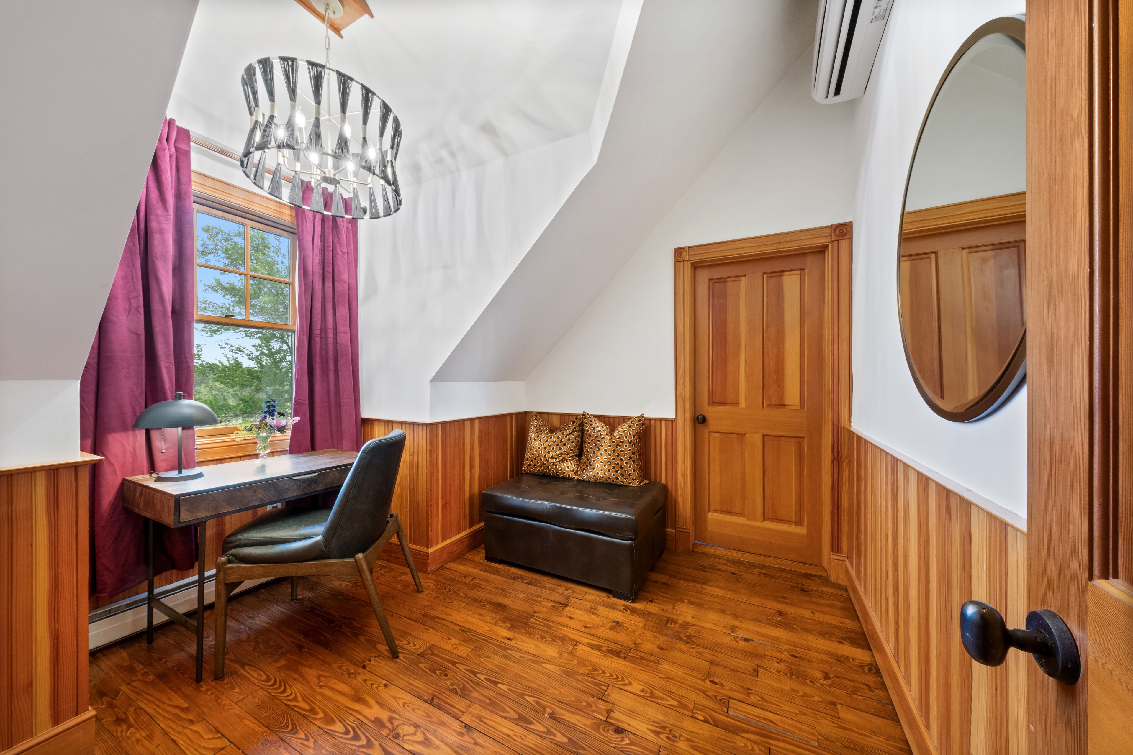 A room with a wooden desk, chair, and bench, a chandelier, a window with curtains, and a door with a round mirror.
