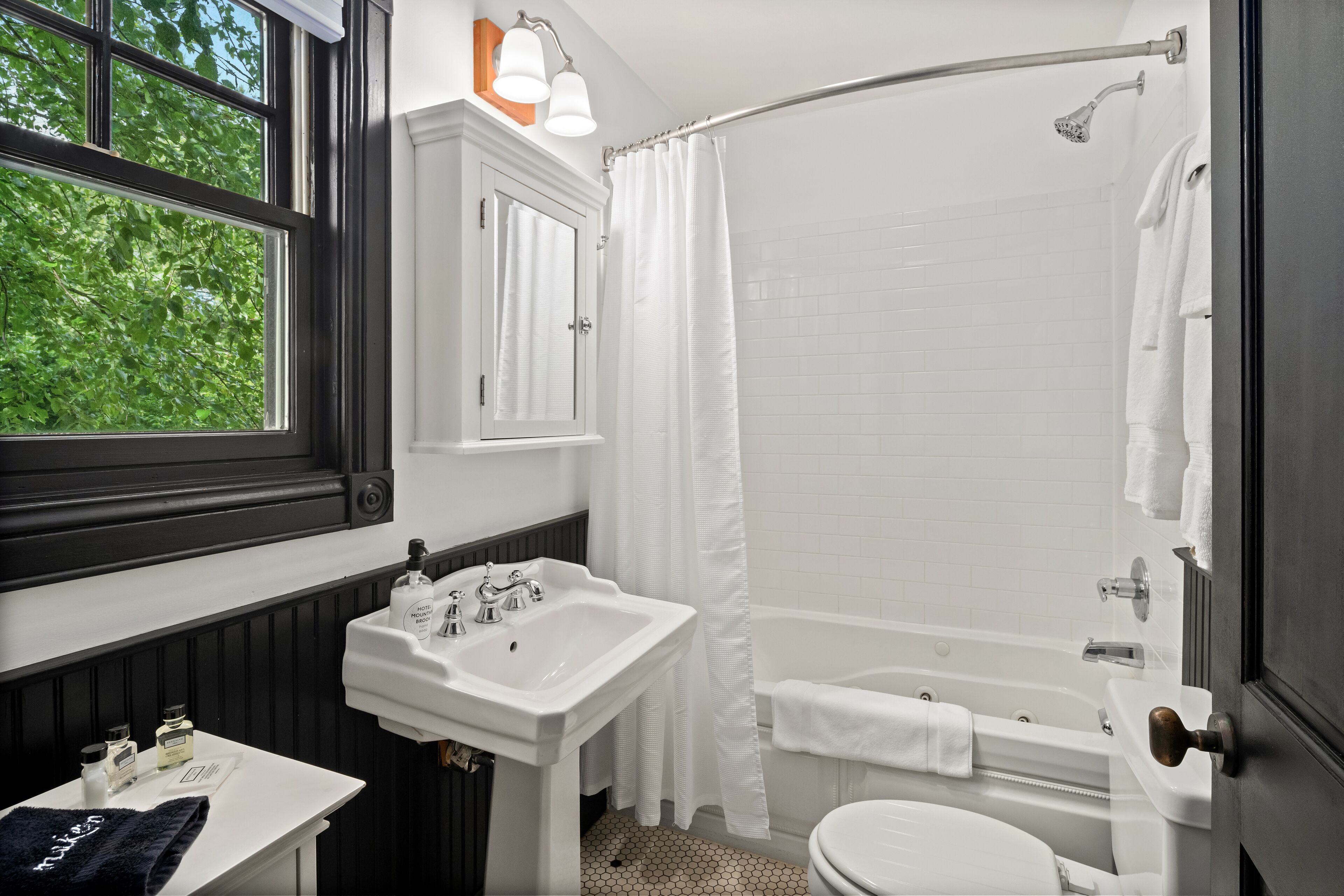 Ein Badezimmer mit weißem Waschbecken, WC und Badewanne. Ein Fenster bietet Blick auf Bäume, und ein Handtuchhalter mit Handtüchern.