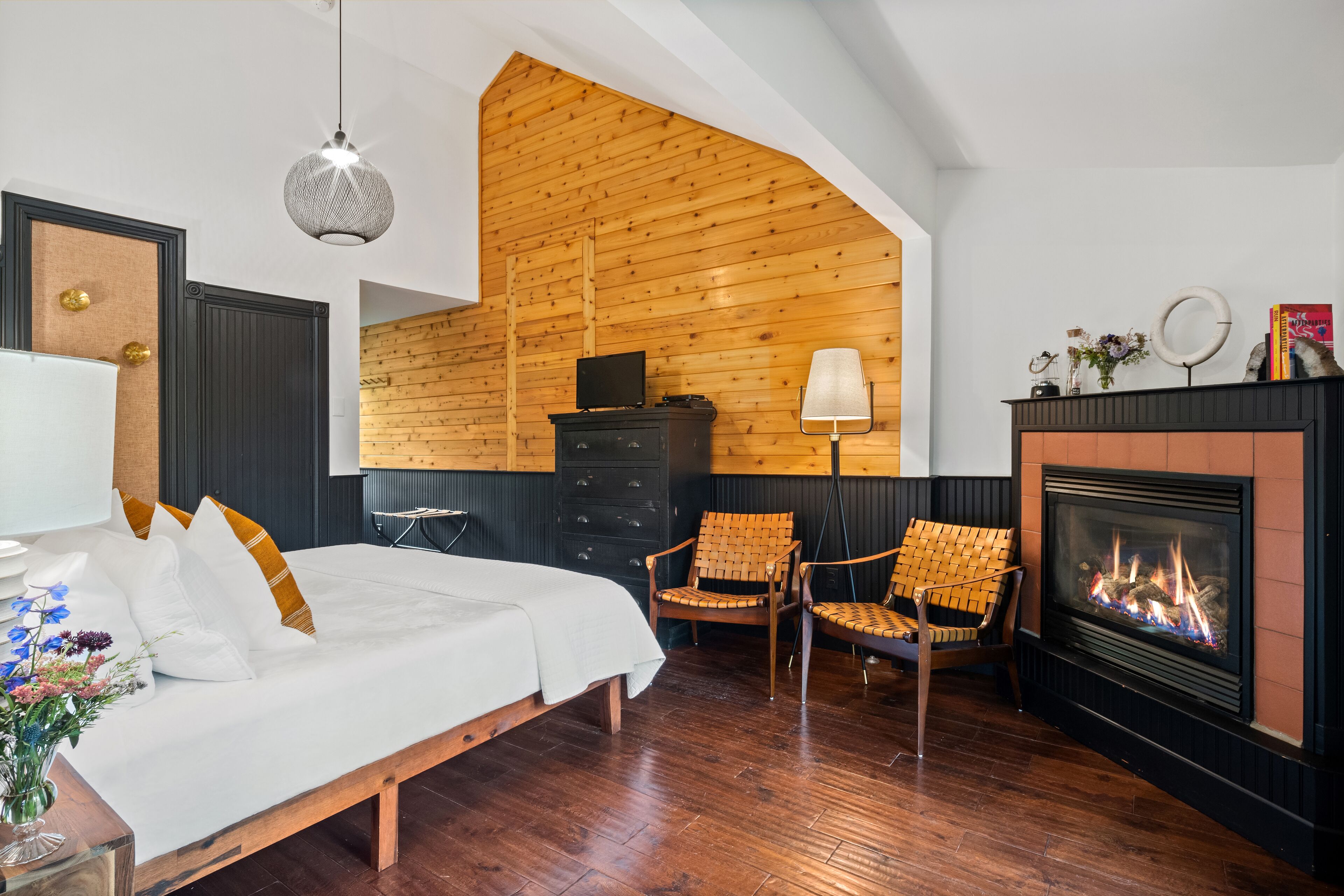 A bedroom with a bed, two chairs, a fireplace, a TV, and a wooden ceiling.
