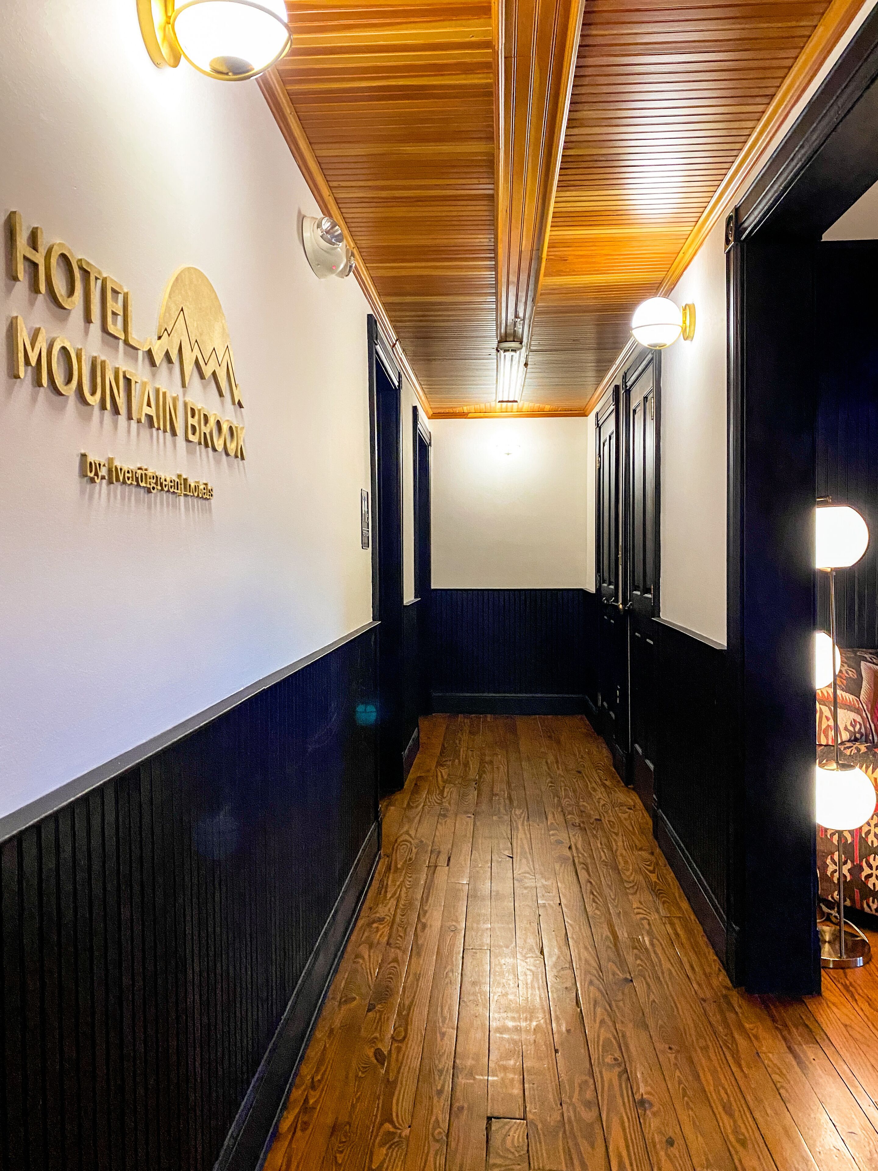 Un couloir d’hôtel avec un plancher en bois, des murs sombres et une enseigne indiquant « HÔTEL MOUNTAIN BROOK, membre du groupe hôtelier Iveragro ».