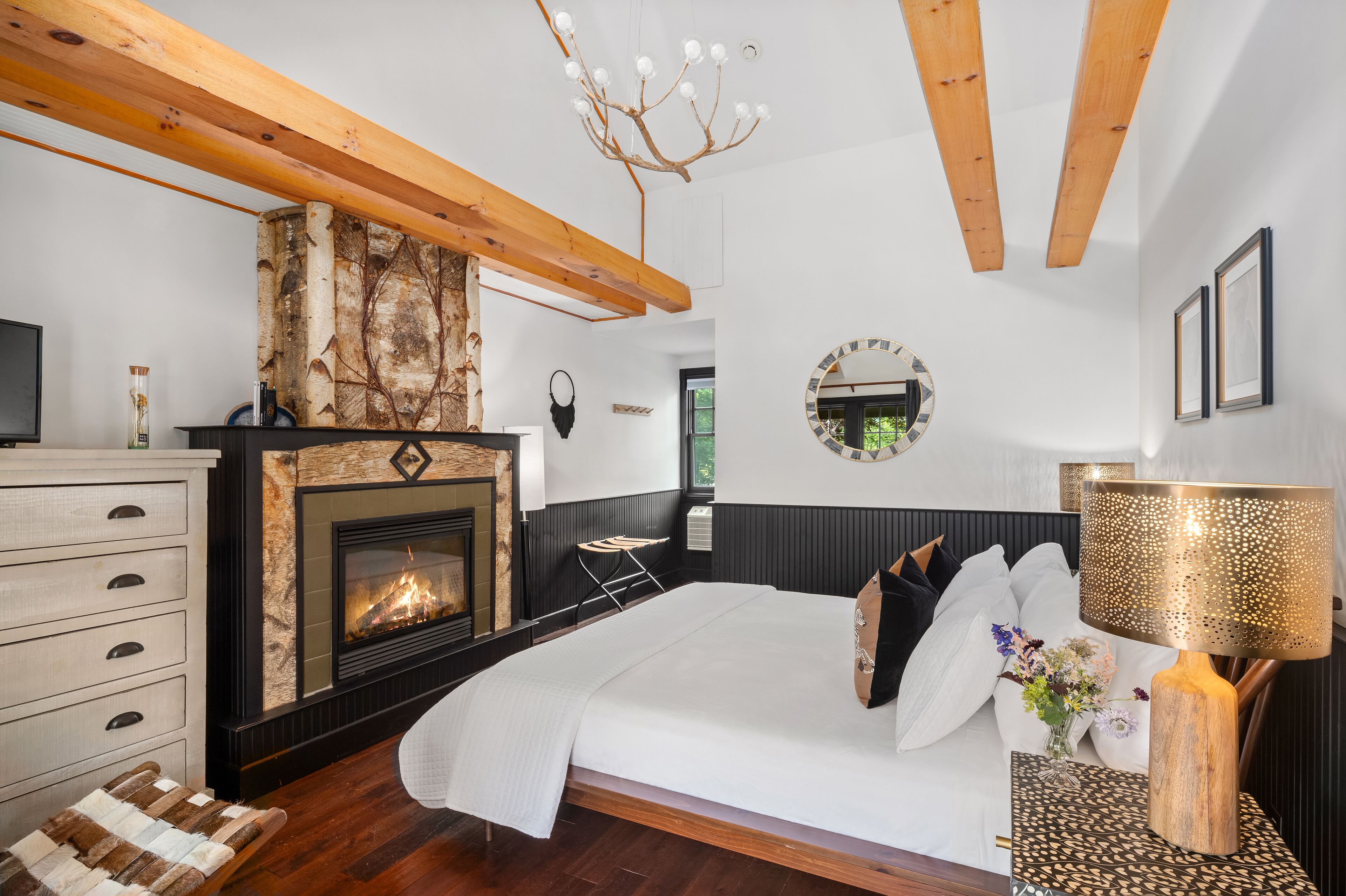 A bedroom with a fireplace, a bed, a nightstand, and a wooden ceiling beam.