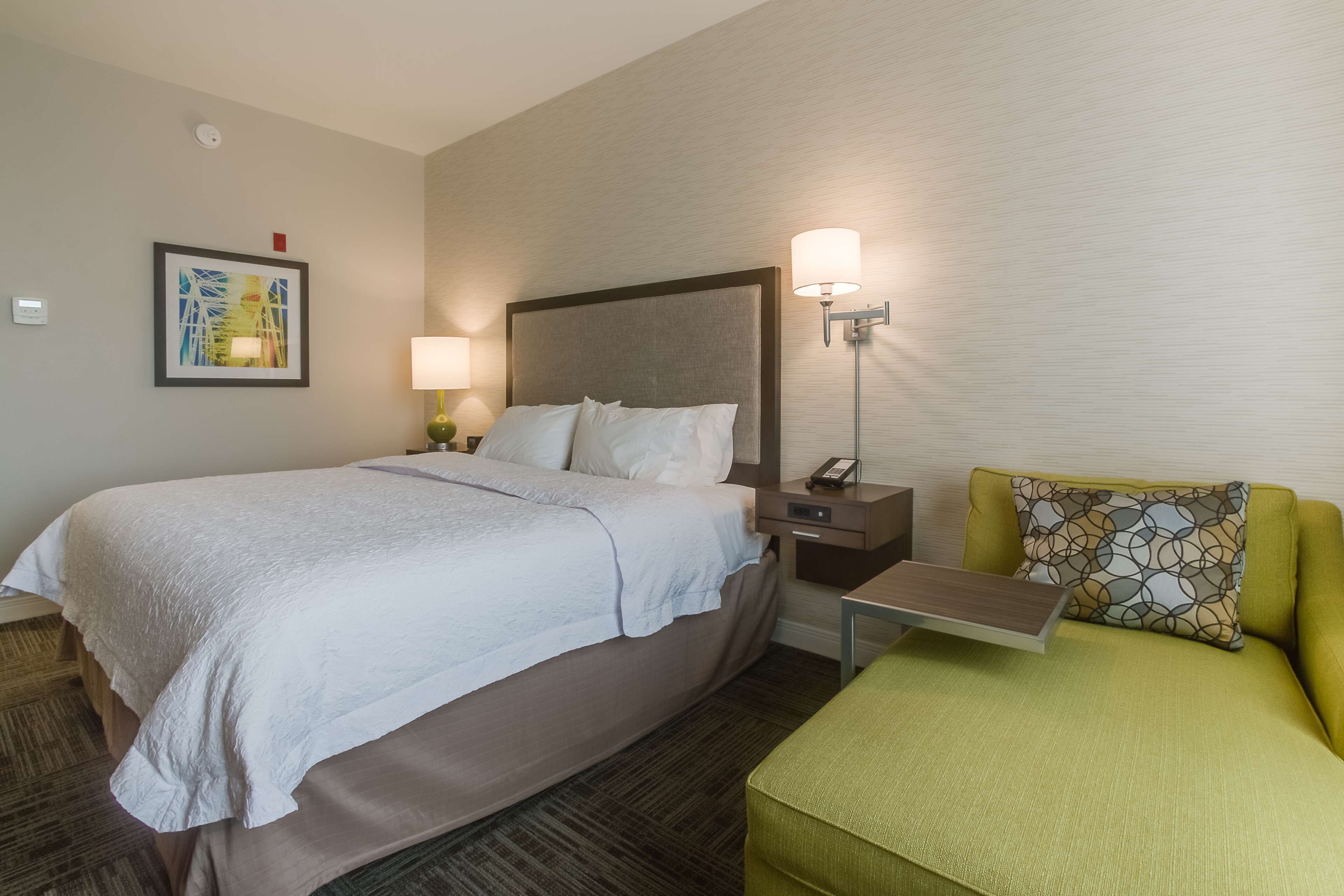 A hotel room with a large bed, a bedside table with a lamp, a small table with a chair, and a sofa with a patterned pillow.
