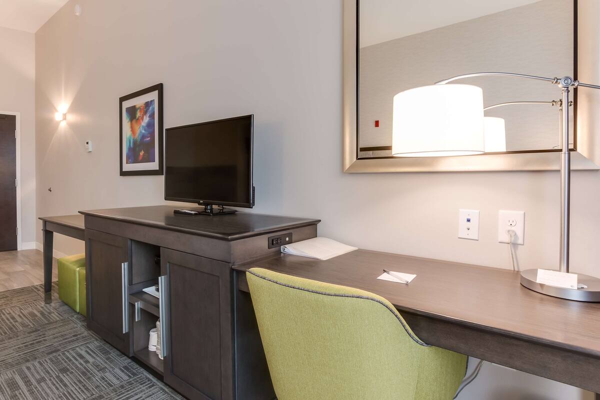 A hotel room with a desk, a television, a mirror, and a green chair.