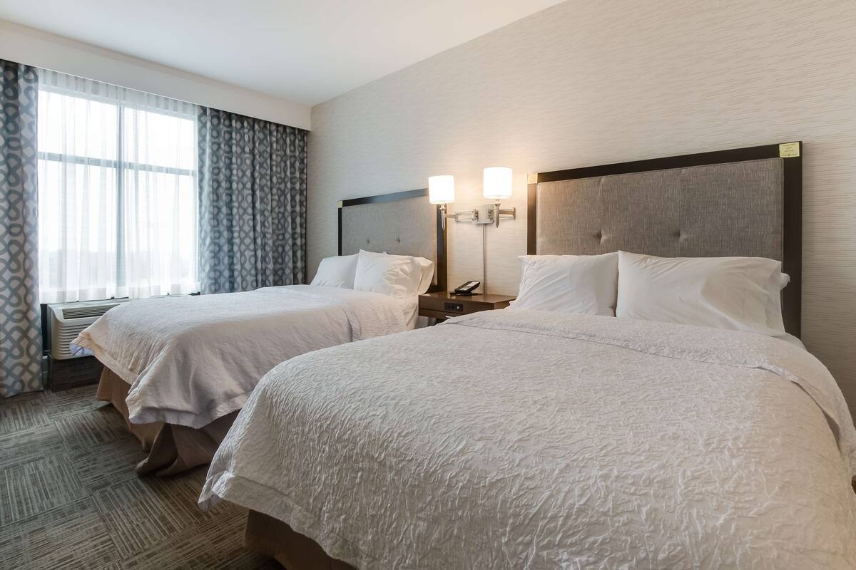 A hotel room with two beds, a bedside table with a lamp, a window with curtains, and a carpeted floor.