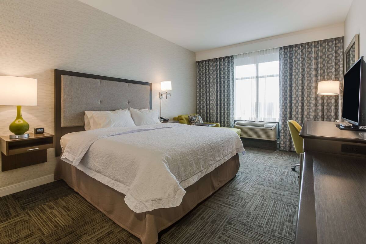 A hotel room with a large bed, bedside tables with lamps, a desk with a TV, a window with curtains, and an air conditioning unit.