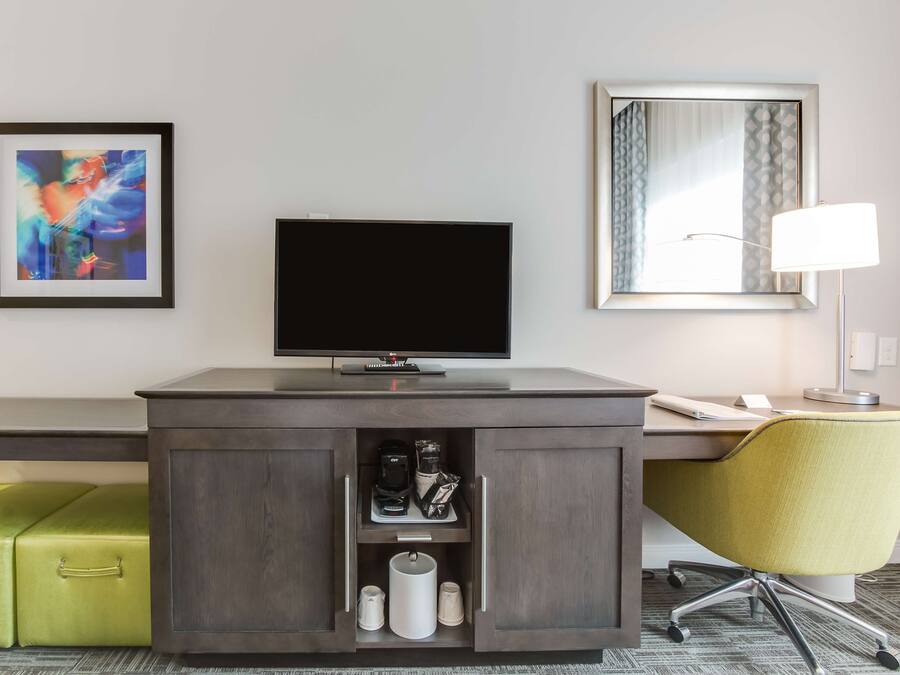 Habitación de hotel con un escritorio de madera oscura, un televisor de pantalla plana, una maleta verde, un sillón amarillo y un cuadro abstracto enmarcado.