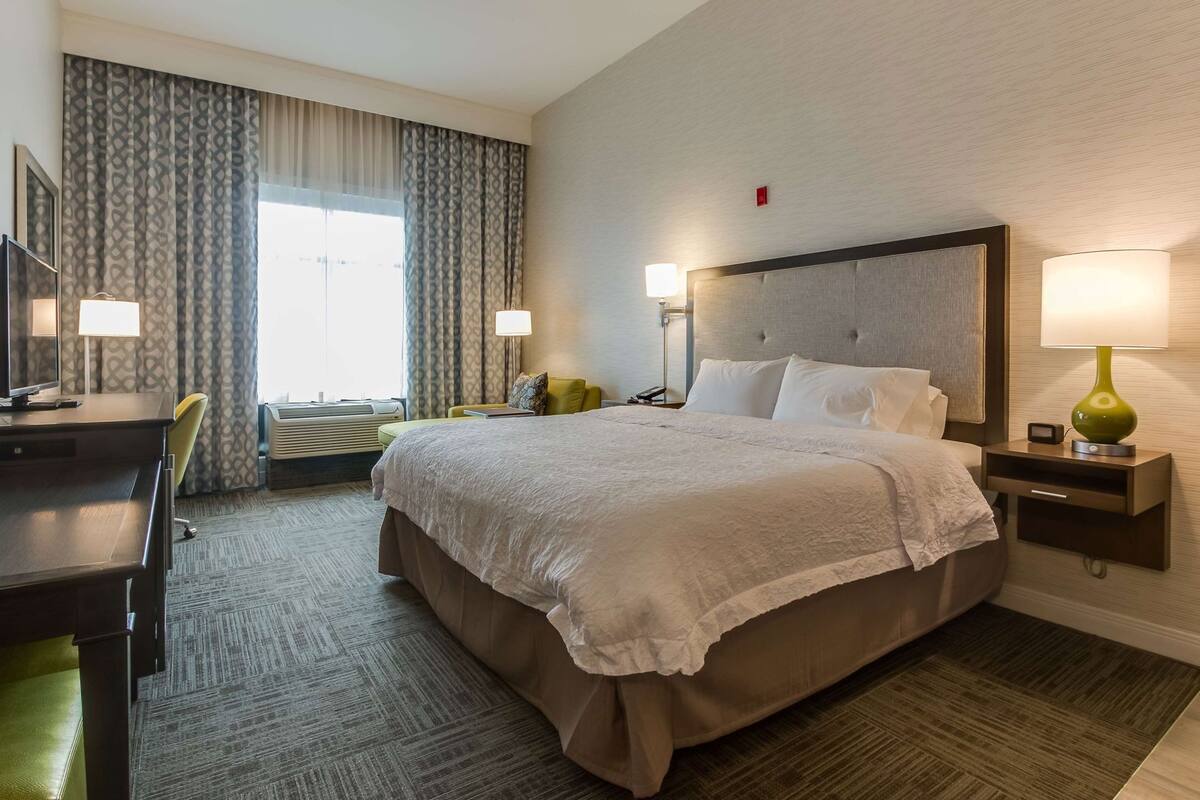 A hotel room with a large bed, bedside lamps, a desk with a chair, and a view of the outdoors through the window.