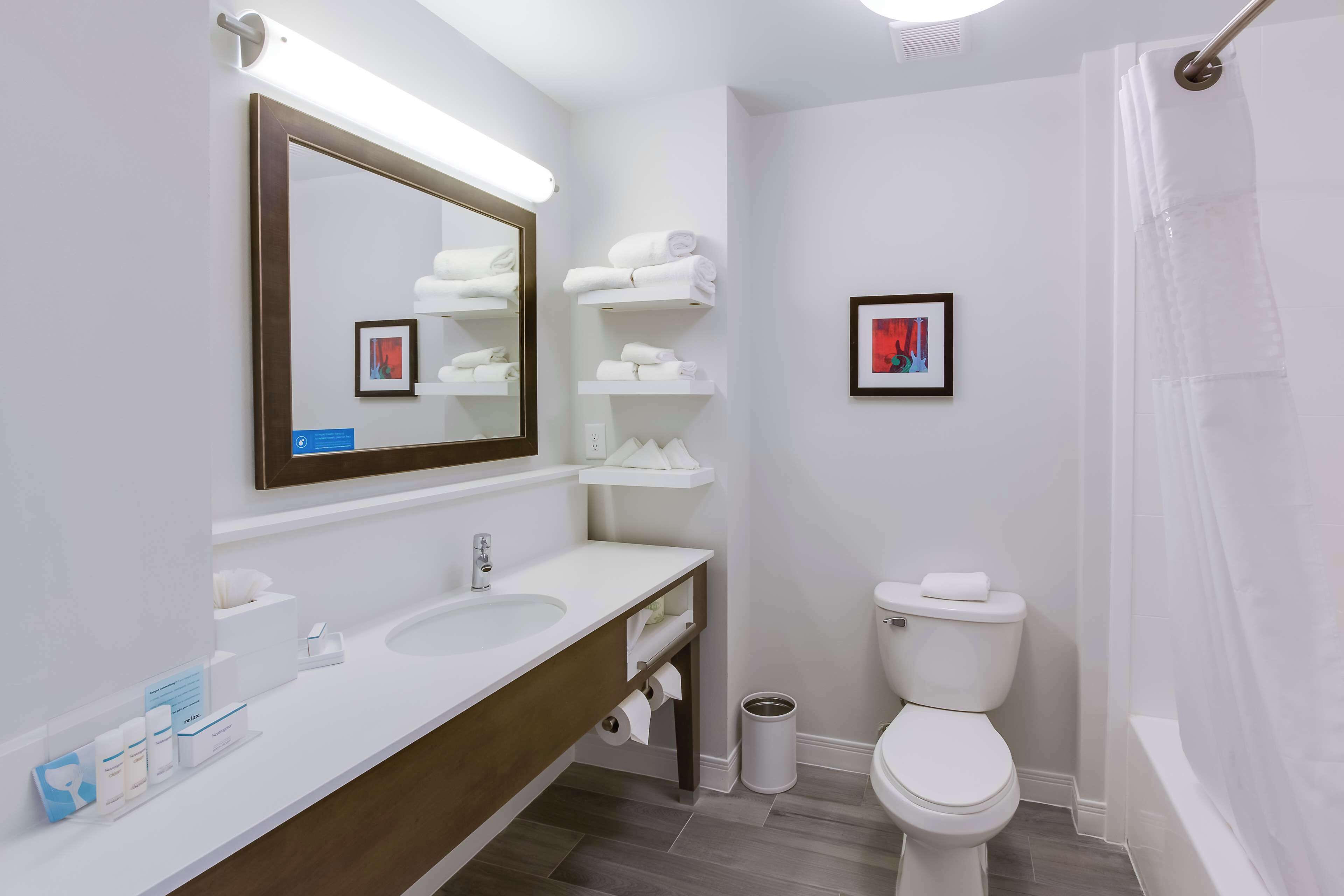 Une salle de bain moderne avec un lavabo blanc, un miroir et des toilettes. On y trouve des étagères avec des serviettes et un tableau encadré accroché au mur.