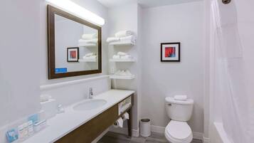 Une salle de bain moderne avec un lavabo blanc, un miroir et des toilettes. On y trouve des étagères avec des serviettes et un tableau encadré accroché au mur.