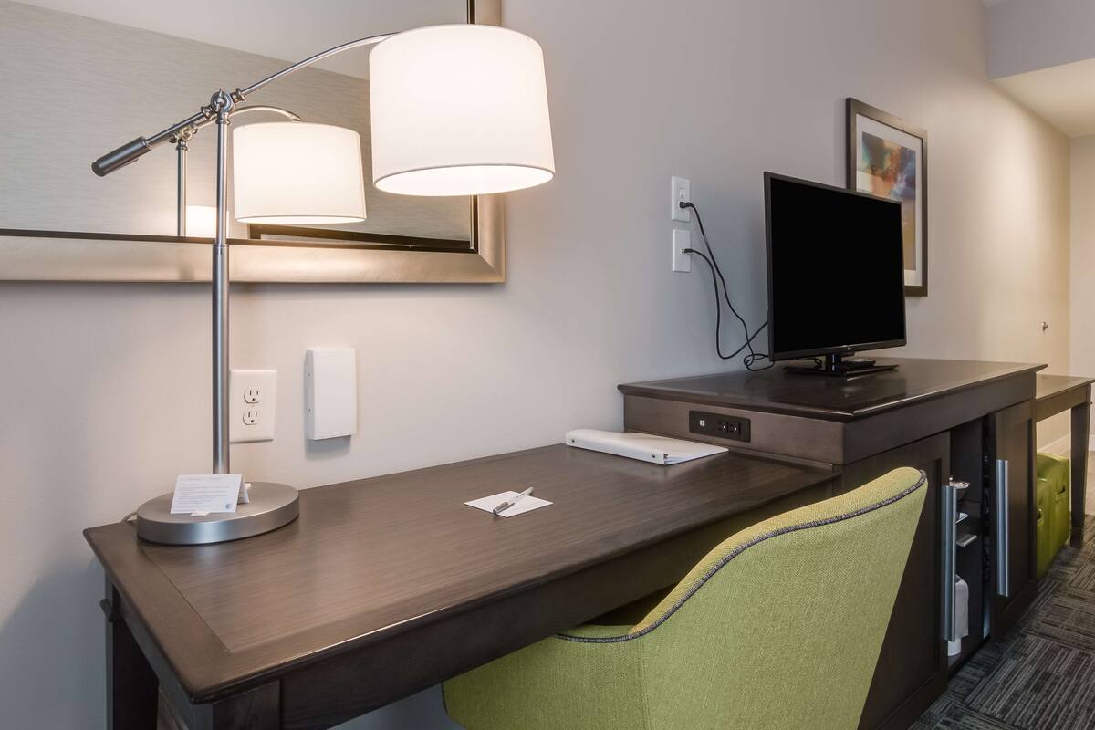 A hotel room with a wooden desk, a lamp, a flat-screen TV, and a green chair.